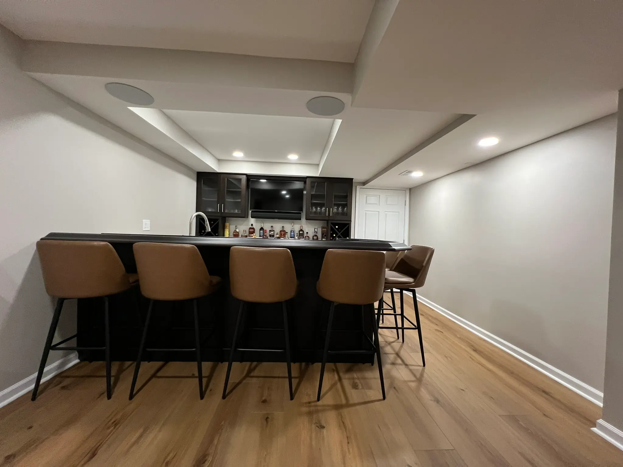 Custom basement wet bar with dark cabinets, leather bar stools, and recessed ceiling speakers