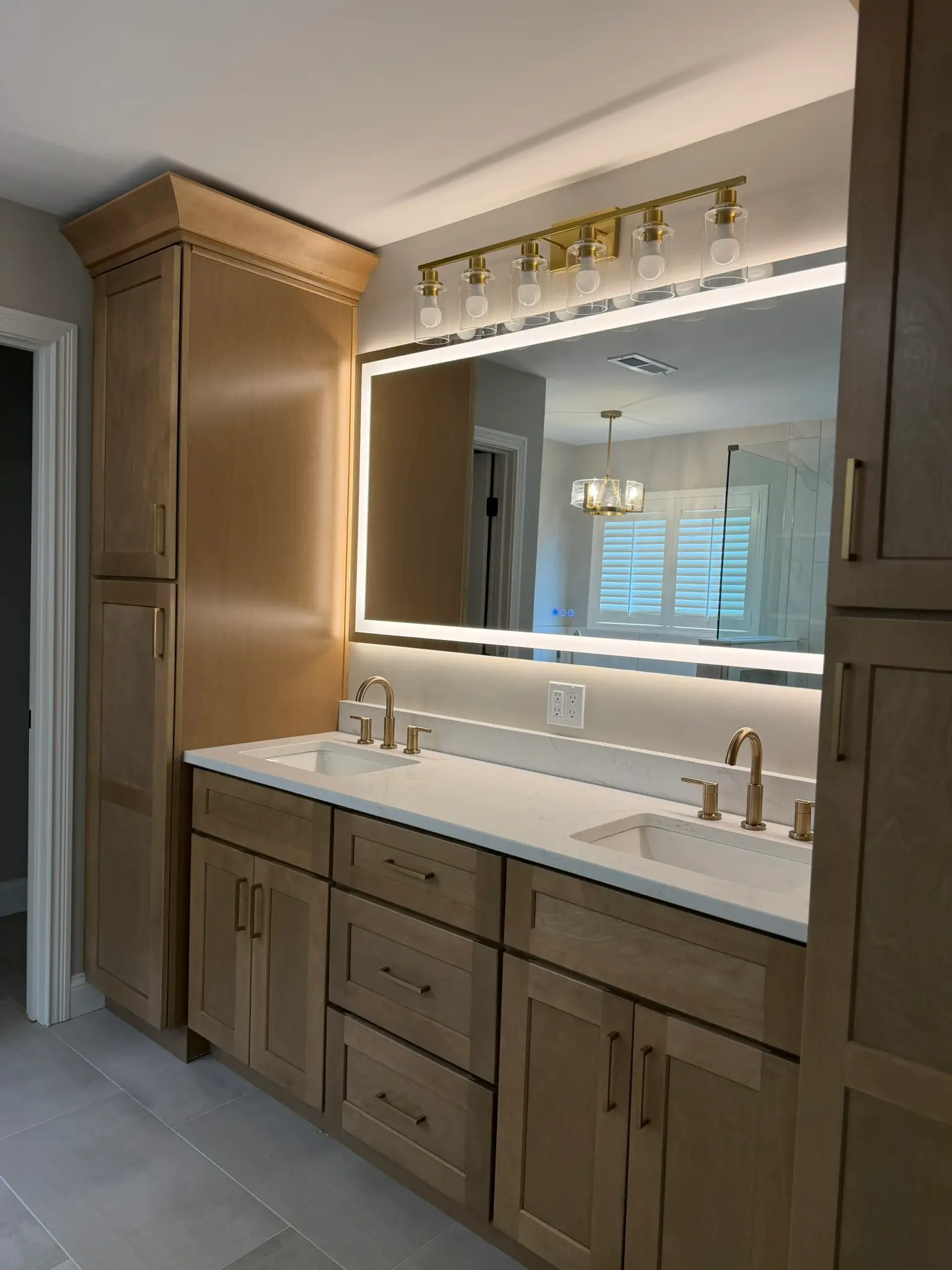 Double vanity with wood cabinetry, matching linen tower, LED backlit mirror, and brass bar lighting