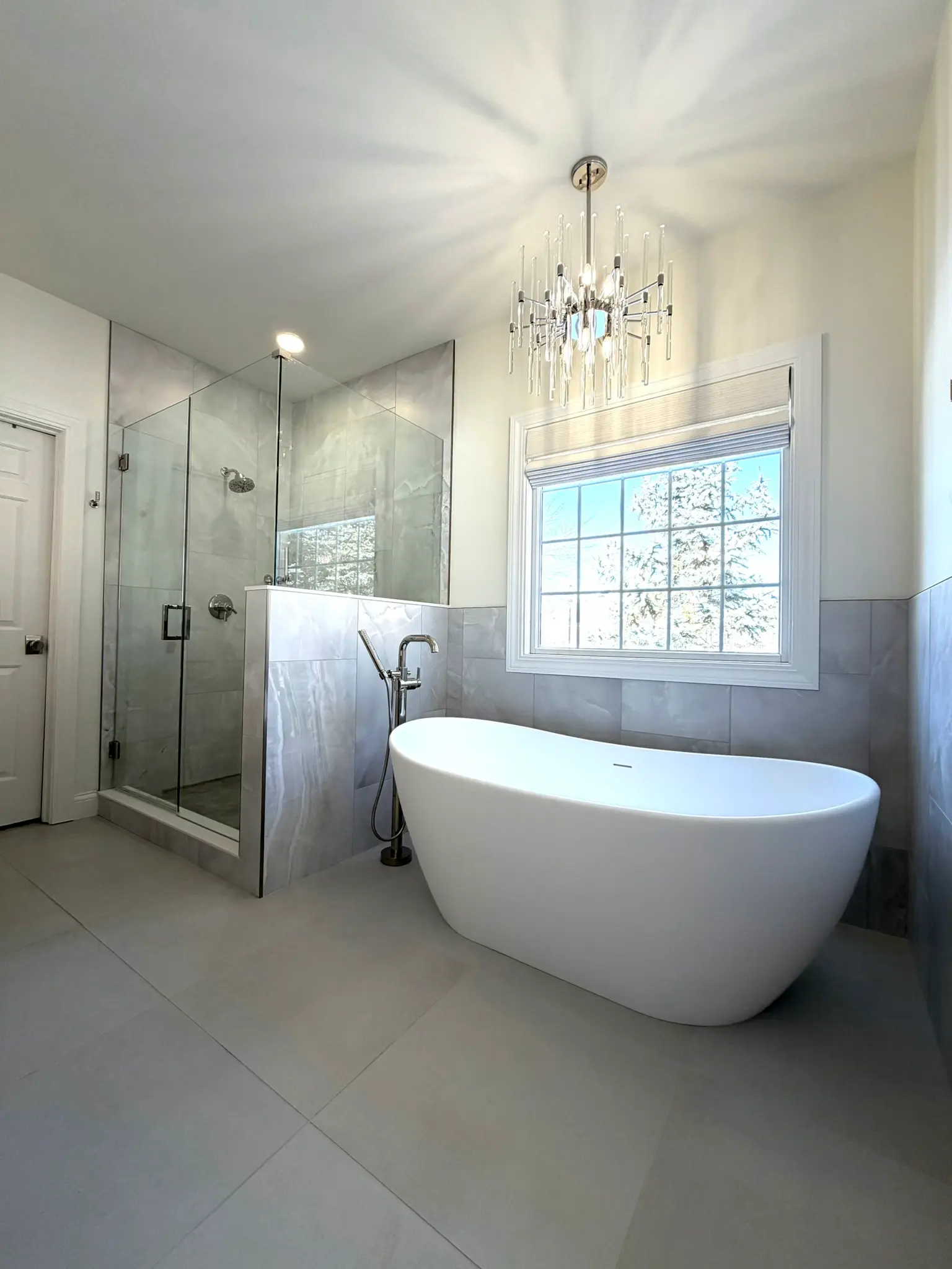 Bright primary suite remodel with freestanding soaking tub, marble tile wall, frameless shower, and crystal chandelier