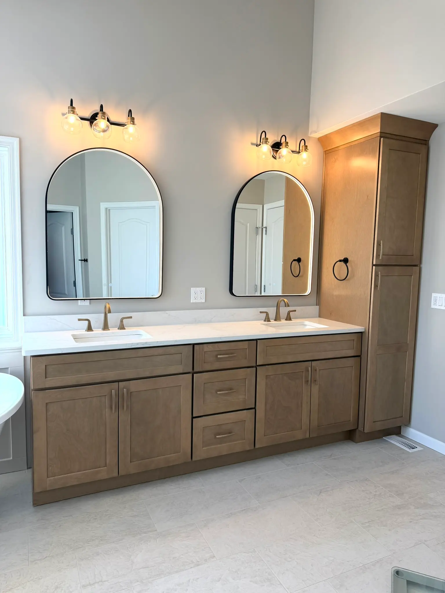 Wide double vanity with natural wood cabinets, linen tower, black arched mirrors, and brass vanity lighting