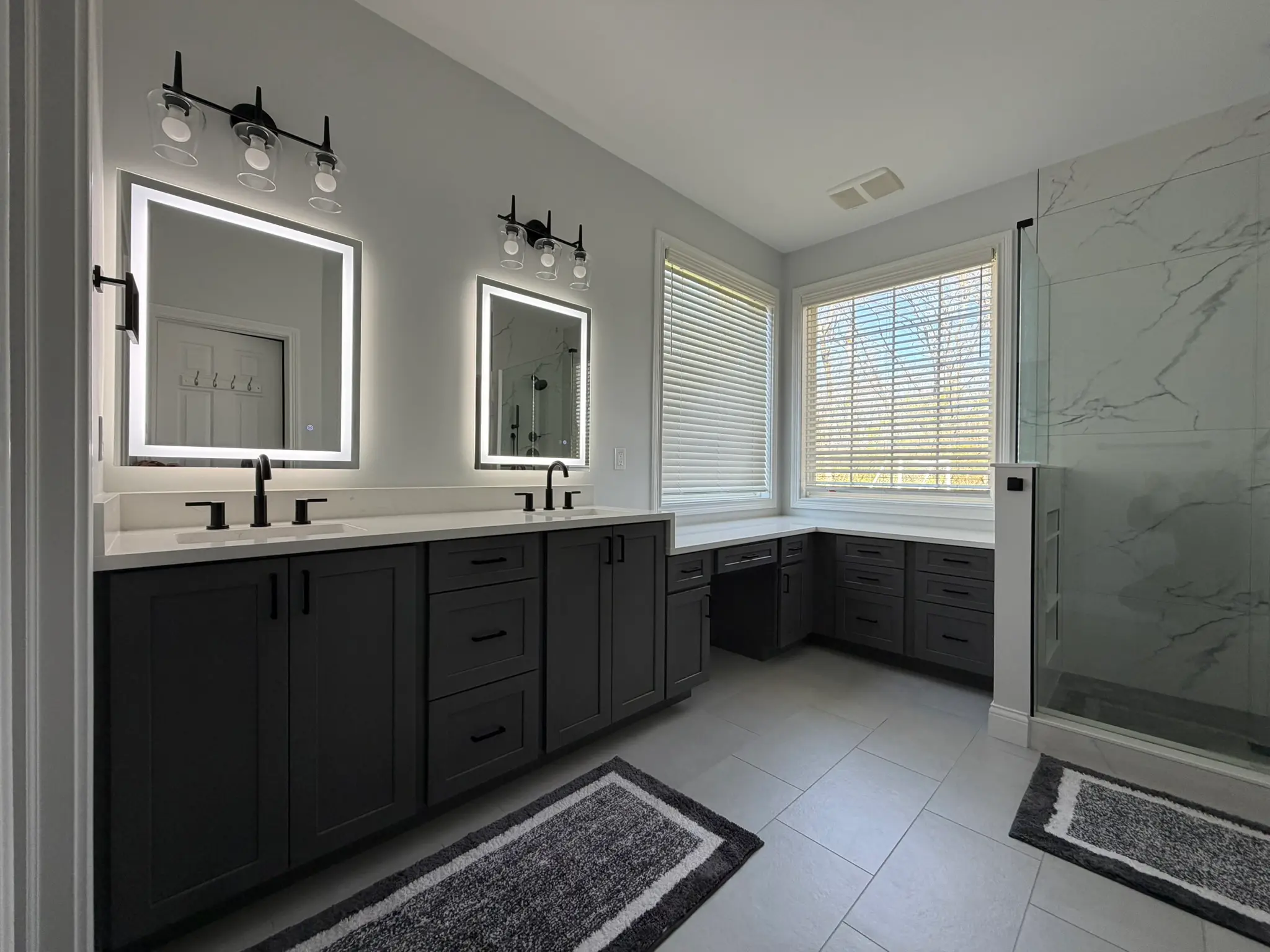 Wide primary bath with charcoal double vanity, makeup area, dual LED mirrors, and marble-look shower