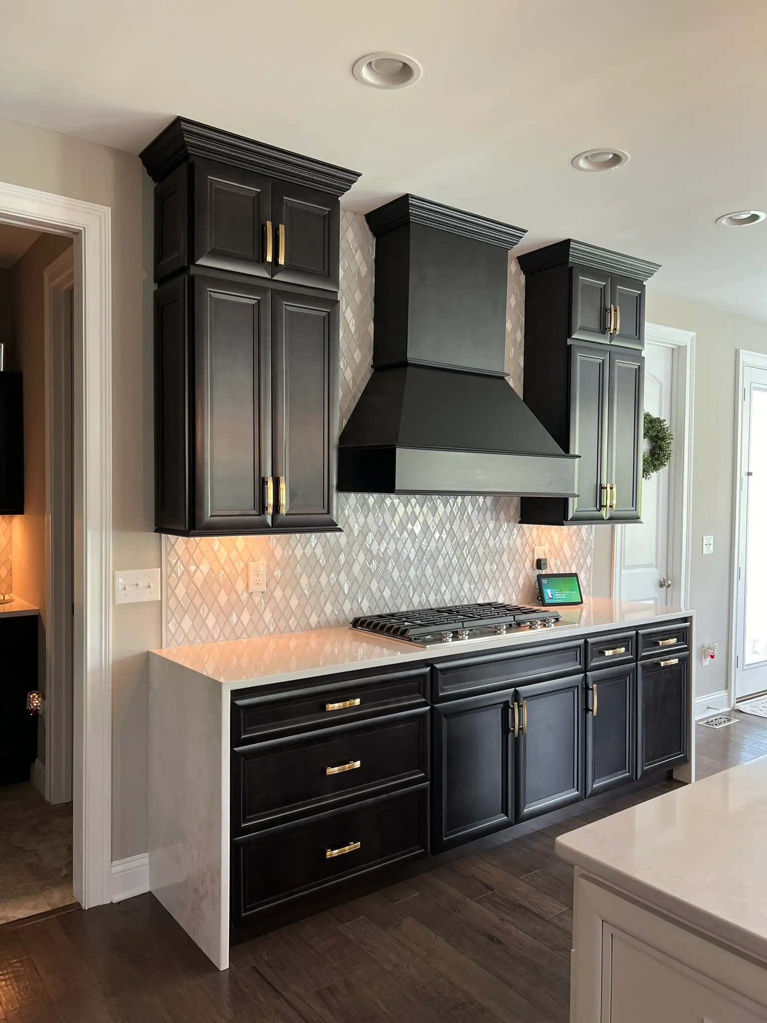 Wide view of black kitchen with custom black range hood, gold hardware, gas cooktop, and pearl diamond tile backsplash