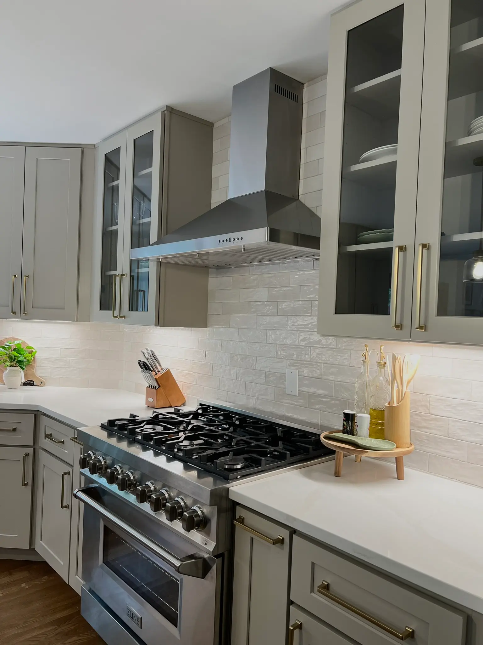 Gray-green kitchen with ZLINE 6-burner gas range, stainless range hood, subway tile backsplash, and glass-front upper cabinets