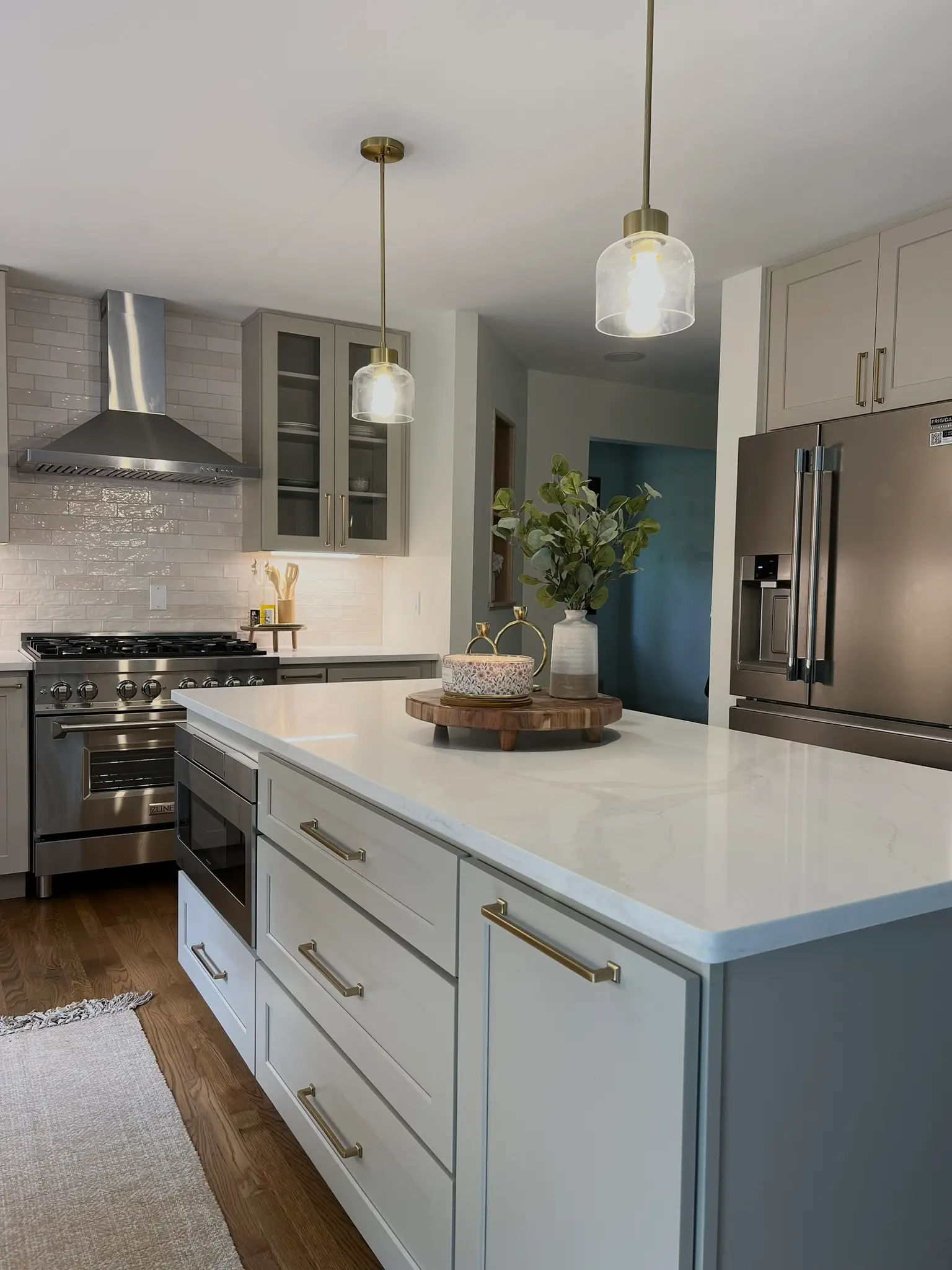 Greige kitchen with white quartz island, glass-front upper cabinets, ZLINE professional range, and brass pendant lights
