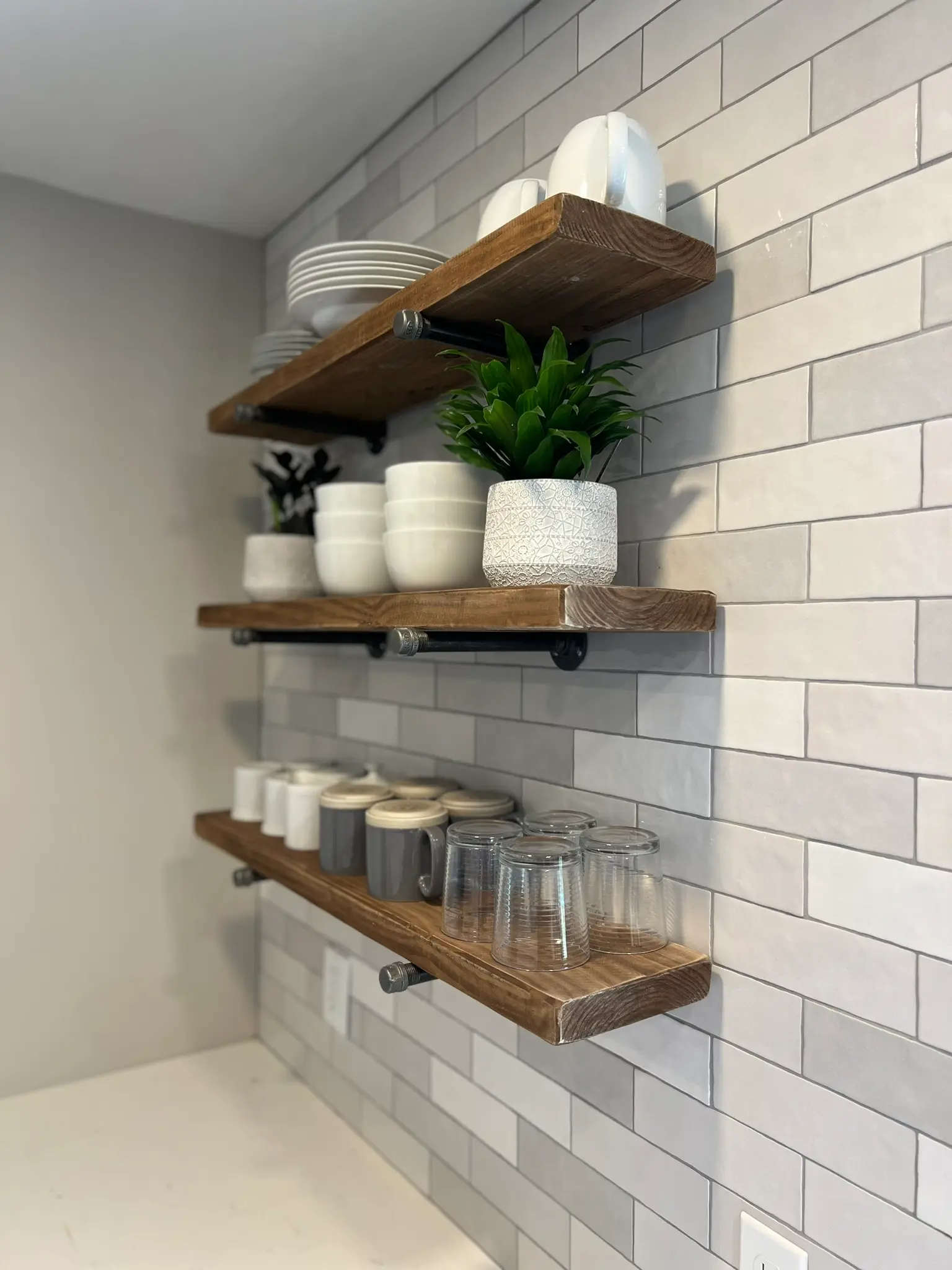 Open floating wood shelves with black pipe brackets on subway tile backsplash, styled with dishware and plants