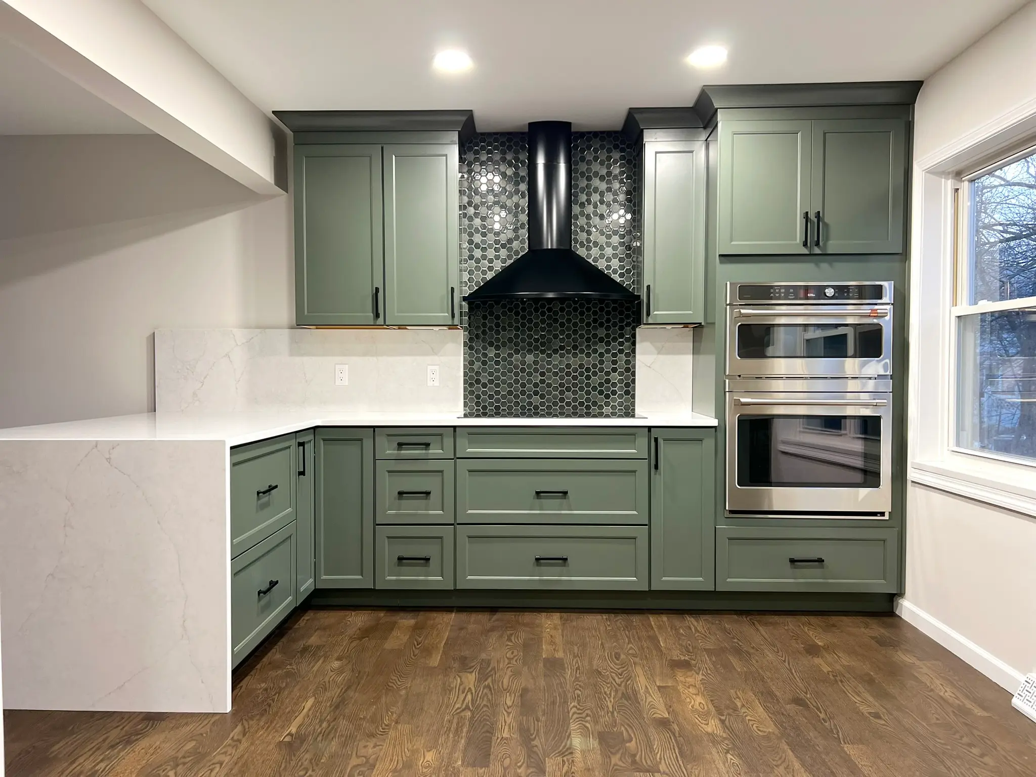 Sage green kitchen with black range hood, hexagon mosaic tile backsplash, white quartz countertop, and built-in double wall oven