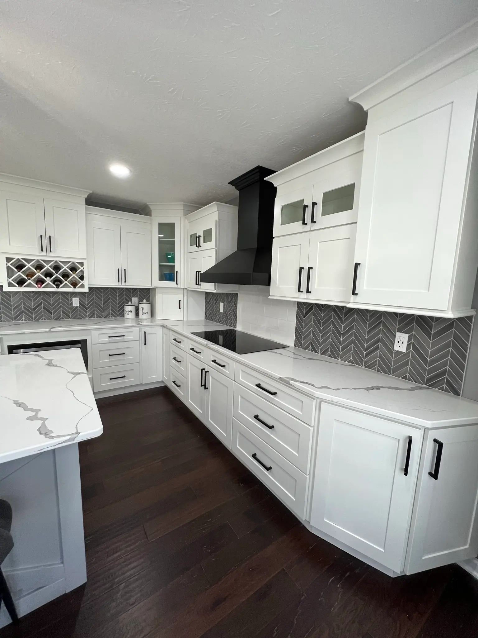 White shaker cabinets with herringbone gray tile backsplash, black range hood, wine rack, and white quartz countertops